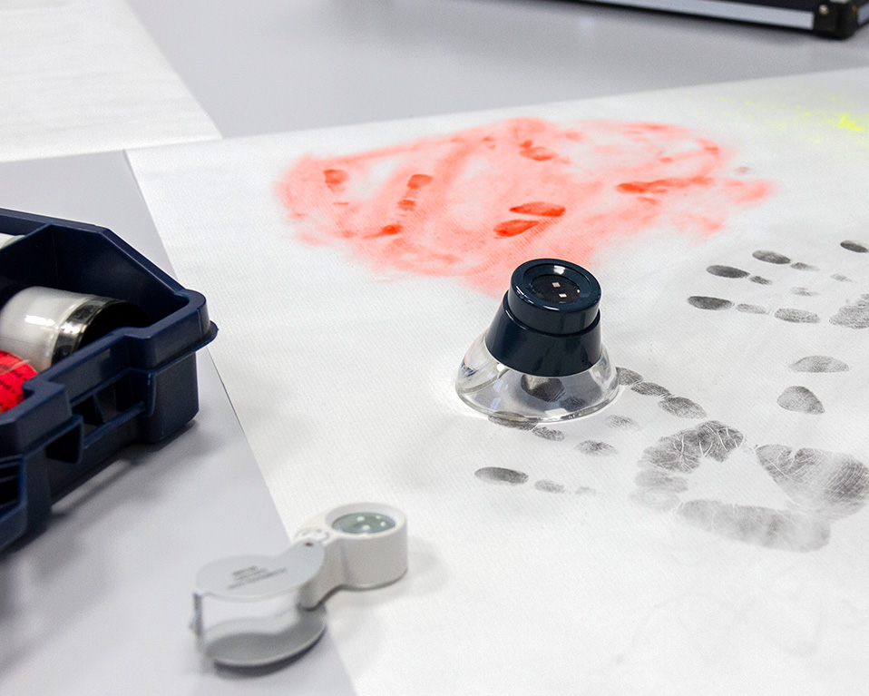 Mesa sobre la que se encuentra un papel que tiene dibujadas varias huellas en varios colores; también se pueden observar distintos instrumentos para el análisis de huellas.